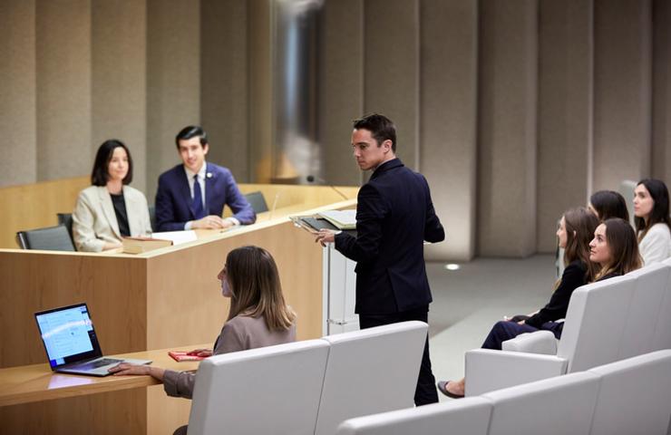 A man is standing and speaking in a courtroom setting, with several people seated around listening.
