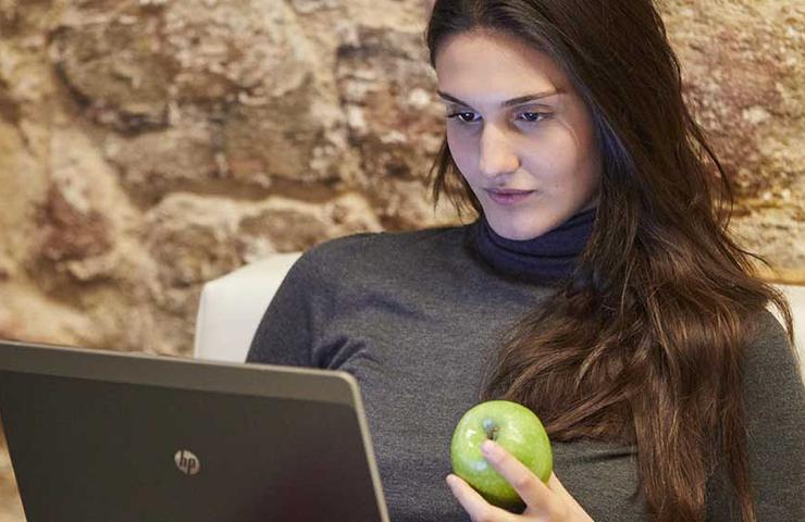 A woman holding a green apple while using a laptop in a cozy indoor setting